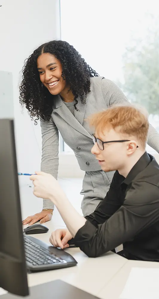 Ein junger Mann sitzt vor einem Computer, eine junge Frau steht lächelnd neben ihm.